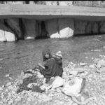 Black and white image of lady carrying baby on her back white getting water at the river to clean clothes
